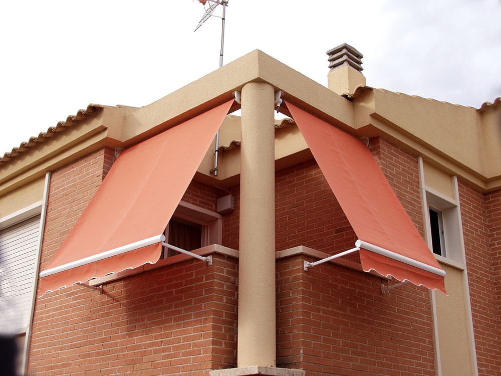 Dos toldos triangulares de color coral instalados en un balcón de una casa de ladrillos, proporcionando sombra.