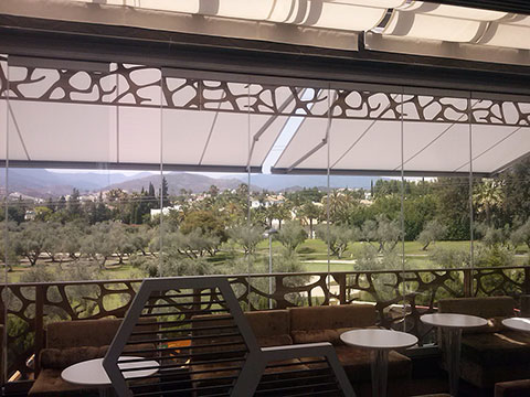 Interior de un restaurante con grandes ventanas protegidas por toldos semitransparentes, ofreciendo una vista panorámica de un paisaje verde.